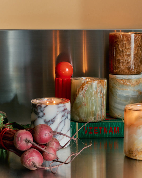 A styled collection of marble and stone candles on a reflective metallic surface, surrounded by decorative props including radishes, a green book, and a copper kettle.
