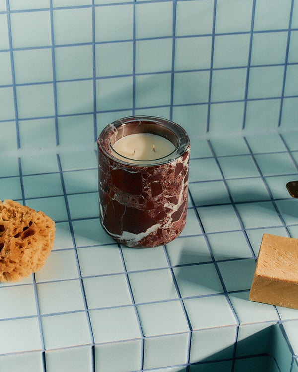 A marble candle holder with a white candle sits on a light blue tiled bathroom counter next to a brass faucet, a natural sponge, and a bar of soap.