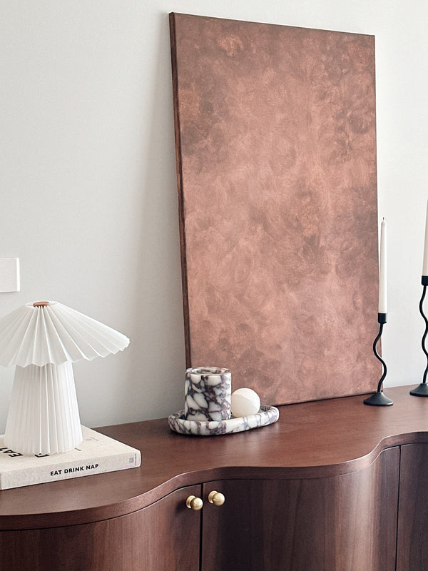 A styled interior scene showing a dark wood sideboard with a large vertical copper-toned panel leaning against the wall, alongside a white pleated table lamp on a book, a small marbled candle holder, and black candlesticks.