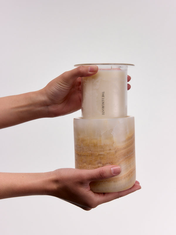 Two hands holding a stack of two translucent beige and tan candles against a plain light gray background. The top candle is slightly offset, showing the brand name printed vertically on its side.